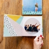 Álbumes de Fotoprix. Foto en uso de un Fotolibro square 21x27 pasándose una página del álbum, se ve también otro cerrado colocados ambos en una mesa.